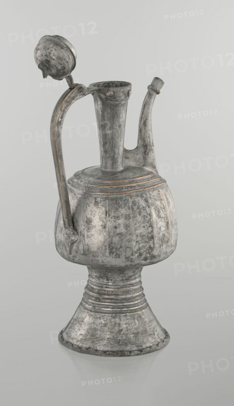 Jug aftabe, Islamic art, jugs, aftabe, water, 19th century, North Caucasus, vessel; height 30.0 cm, length 14.8 cm, diameter 12.0 cm, A metal pitcher with a rounded body, narrow neck, curved spout, and tall flared base sits against a pale background; the surface shows a weathered, textured finish with areas of darker gray, lighter gray, and hints of muted brownish tones; a curved handle rises from the body to the neck and ends near a hinged, domed lid that is open, the lid sharing the same worn gray and brown coloration; the body has subtle vertical ridges and horizontal bands near the top, and the flared pedestal base features concentric ridges and a scalloped edge, all reflecting the same aged gray and brown color palette.