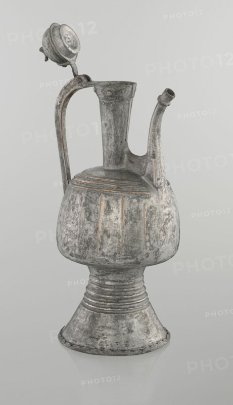 Jug aftabe, unknown provenance; Islamic art; jug used for water aftabe; 19th century; North Caucasus; vessel; height 30.0 cm, length 14.8 cm, diameter 12.0 cm, A metal vessel with a tall narrow neck, a curved spout, and a looped handle topped by a hinged lid, all showing a worn, textured surface. The body of the vessel is bulbous above a flared pedestal base with horizontal ridges where the body meets the stem. Vertical incised grooves decorate the rounded midsection, and concentric incised bands appear near the shoulder. The hinged lid sits open and is circular with a domed top. The spout is slightly curved and tapers toward its opening. The surface displays a mottled, aged appearance with visible areas of abrasion and patina. The visible colors are shades of gray, silver, off-white, and touches of muted brown or rust. The background is a smooth, light gray.