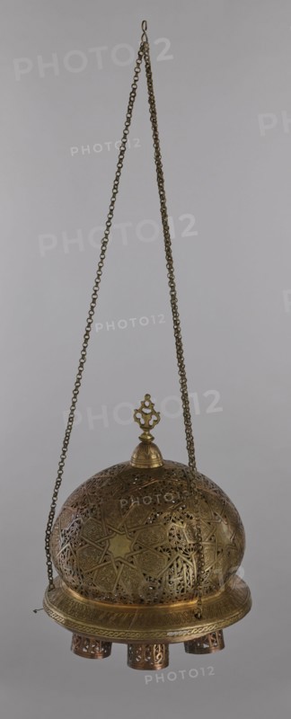 Lamp, Islamic art, Arabic culture; metal lighting fixture with geometric ornament and Arabic inscriptions. Cover decorated with a star motif. Disc base with seven holes for glass sleeves glass oil containers missing. 19th–20th century, Damascus, Syria. Lamp; height 40 cm, diameter 32 cm, weight 2.4 kg., A hanging metal lantern suspended by a long decorative chain, with a hook at the top and a pendant finial above a domed, perforated body; the lantern features intricate geometric and floral openwork patterns across the dome and band, and a circular base with short cylindrical feet that also have cutout designs. The visible colors are shades of brass and bronze, with warm golden brown metallic tones and subtle darker patina in the recessed areas, set against a plain light gray background.