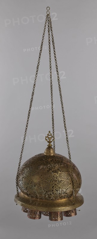 Lamp, Islamic art, Arabic culture; metal lamp with Arabic inscriptions and geometric ornament including a star motif on the cover. Disc base with seven holes for glass sleeves glass oil containers missing. 19th–20th century, Damascus, Syria. Height 40 cm, diameter 32 cm, weight 2.4 kg., A hanging metal lantern suspended by a group of chains, with a hooked loop at the top and three chains descending to a decorative finial; the lantern body is a domed, perforated and intricately patterned structure with geometric and floral openwork, showing metallic tones of brass and bronze with areas of darker patina and hints of copper; a circular rim supports a few short cylindrical pendants with pierced patterns beneath the dome; the background is a plain neutral gray.