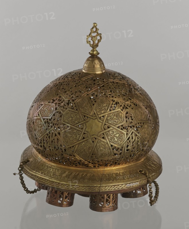 Lamp, creator unknown, Islamic art, Arabic culture; metal oil lamp with geometric ornament and Arabic inscriptions; cover decorated with a star motif; disc base with seven holes for glass sleeves glass oil containers missing. 19th–20th century, Damascus, Syria. Height 40 cm; diameter 32 cm; weight 2.4 kg., A rounded, ornate metal object with a domed lattice upper section and a circular base, primarily gold and brass in color with darker bronze accents; the dome features intricate geometric and floral cutwork with a star-shaped motif and repeating patterned panels, and a decorative finial sits atop a small rounded cap; the base has engraved banding and a braided decorative rim with a few hanging chain pieces, and the whole piece rests on a group of short cylindrical feet with pierced patterns, all set against a neutral light gray background.