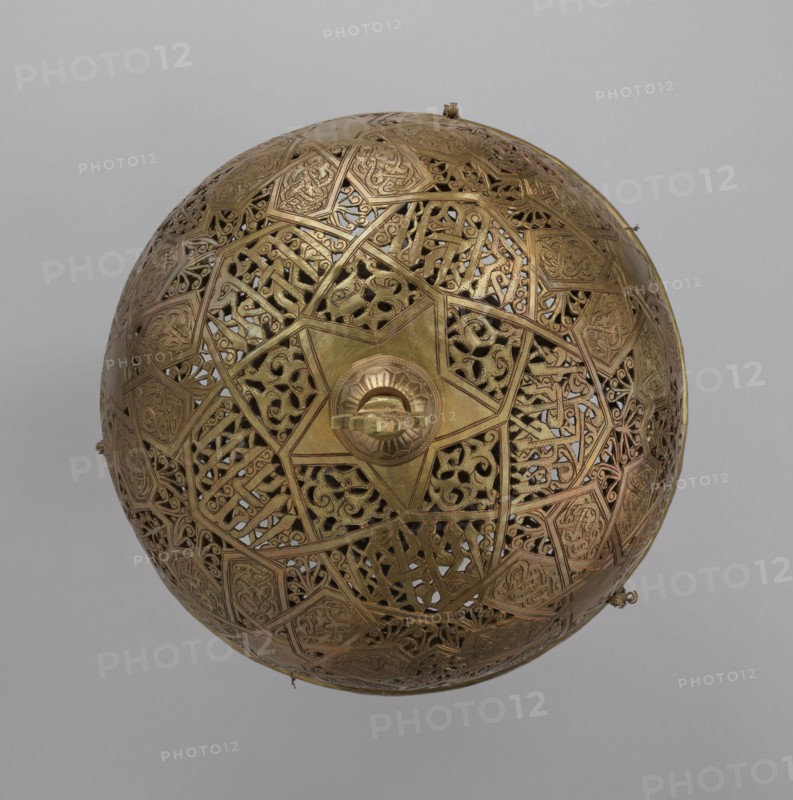 Lamp, 19th–20th century, Damascus, Syria. Metal lighting lamp with Arabic inscriptions and geometric ornament; cover decorated with a star motif. Disc base with seven holes for glass sleeves glass oil containers missing. Height 40 cm; diameter 32 cm; weight 2.4 kg., A spherical metal object with an ornate openwork surface made of interlocking geometric panels and filigree patterns, primarily brass-colored with areas of darker and lighter golden tones; a central raised medallion with a circular handle sits amid star-shaped motifs, surrounded by intricate latticework and curving decorative forms; the background is a uniform light gray.