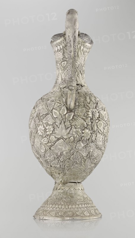 Jug, purchase, Islamic art, jugs, zoomorphism, coats of arms, floral motifs, plant ornament, leaves, carnations, plant tendrils. A tall silver jug with a pear-shaped base. The tall, conical base tapers upward, smoothly merging into a wide spout. The entire vessel is richly decorated with plant motifs—leaves, twigs, and various species of flowers. In the center of the belly wall, on one side only, is a blank shield. The jug's handle is decorated with images of spotted animals with large ears and open mouths. A hinge at the upper edge of the handle indicates the vessel originally had a lid. Late 19th century, possibly Northern India or Iran; height 43 cm, weight 1795 g., An ornate metal vessel with a long, curved neck and bulbous body sits upright on a flared base, all covered in dense raised floral and leaf patterns; the surface shows intricate carved motifs, textured bands, and a sculpted spout that projects from the neck area, with a small decorative element beneath it; the piece appe...