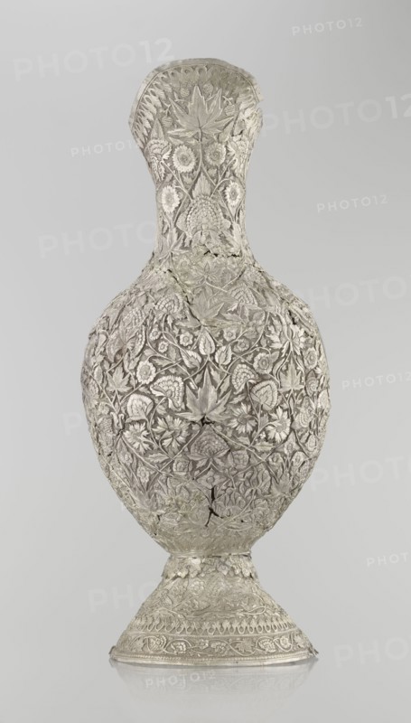 Jug, late 19th century, Northern India, or Iran, silver vessel, height 43 cm, weight 1795 g. A tall silver jug with a pear-shaped body and a tall conical neck that tapers upward into a wide spout. The entire surface is richly decorated with plant motifs—leaves, twigs, various flowers including carnations, and plant tendrils. On one side of the belly is a blank shield coat of arms. The handle is ornamented with zoomorphic figures: spotted animals with large ears and open mouths. A hinge at the upper edge of the handle indicates the vessel originally had a lid., A tall ornate vessel with a narrow neck and rounded body standing on a flared foot, entirely covered in intricate raised floral and leaf patterns, with visible seams and a few cracks in the surface; the surface appears metallic with shades of silver and pale gray, and the background is a smooth light gray.