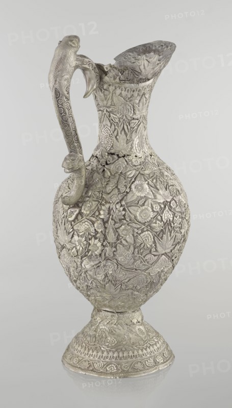 Jug, late 19th century, Northern India, or Iran, silver vessel, height 43 cm, weight 1795 g. A tall silver jug with a pear-shaped body and a tall, conical neck that tapers upward into a wide spout. The entire surface is richly chased with plant motifs—leaves, twigs, tendrils, carnations and other flowers. On one side of the belly is a blank shield. The handle is decorated with spotted animals with large ears and open mouths; a hinge at the upper edge of the handle indicates the jug originally had a lid. Style: Islamic art; motifs: zoomorphism, coats of arms, floral ornament., A single ornate metal pitcher with a curved handle and a flared spout is shown against a plain light gray background; the pitcher has an overall silvery-gray color with a slightly darker patina in recessed areas and is covered in dense raised floral and leaf patterns, including stylized blossoms, vines, and textured foliage across the body, neck, and base, while the handle features repeating decorative motifs and ...