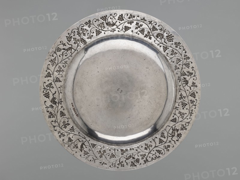 Platter, purchased provenance, Chinese art, Chinese culture, Qing 1644–1911, grapes, decorative vessel, squirrels, vines, 19th–20th century, China, vessel, weight 1170 g, diameter 35.0 cm, A round metallic plate with a broad decorative rim and a slightly recessed central well, showing a smooth reflective surface and visible scratches and wear; the rim features an ornate vine-and-grape pattern of engraved curling stems, leaves, and clusters arranged around the circumference; the metal appears silver-gray with darker gray accents in the engraved areas, and the background is a uniform muted gray.