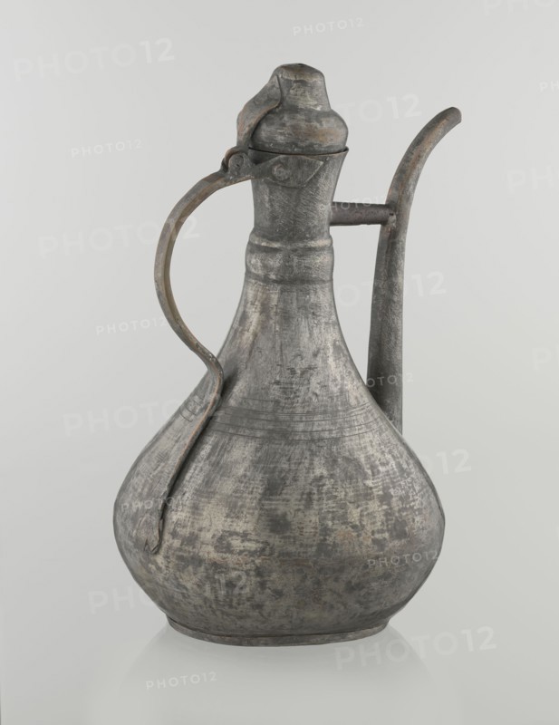 Jug aftabe, Islamic art, Islamic culture, jugs, water, geometric ornament, 19th century, Caucasus, Central Asia, vessel; height 31.5 cm, width 20.5 cm, diameter 18 cm, A metal pitcher with a bulbous body, narrow neck, and hinged lid sits against a light background; it has a curved handle attached near the neck and a long, slightly curved spout on the opposite side, showing a weathered, textured surface with visible wear and patina in shades of gray, silver, dark gray, and hints of brown.