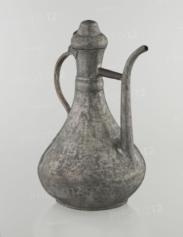 Jug aftabe, Islamic art, Islamic culture, jugs, water, geometric ornament, 19th century, Caucasus, Central Asia, vessel; height 31.5 cm, width 20.5 cm, diameter 18 cm, A metal pitcher with a bulbous base and a narrow neck sits against a pale background, showing a weathered surface with mottled shades of gray and patches of darker and lighter gray, along with small areas of muted brownish and rusty tones; it has a curved handle attached near the neck and a long curved spout extending from the shoulder, both sharing the same textured, aged gray appearance, and a fitted lid with a rounded top that matches the overall metallic, worn finish.