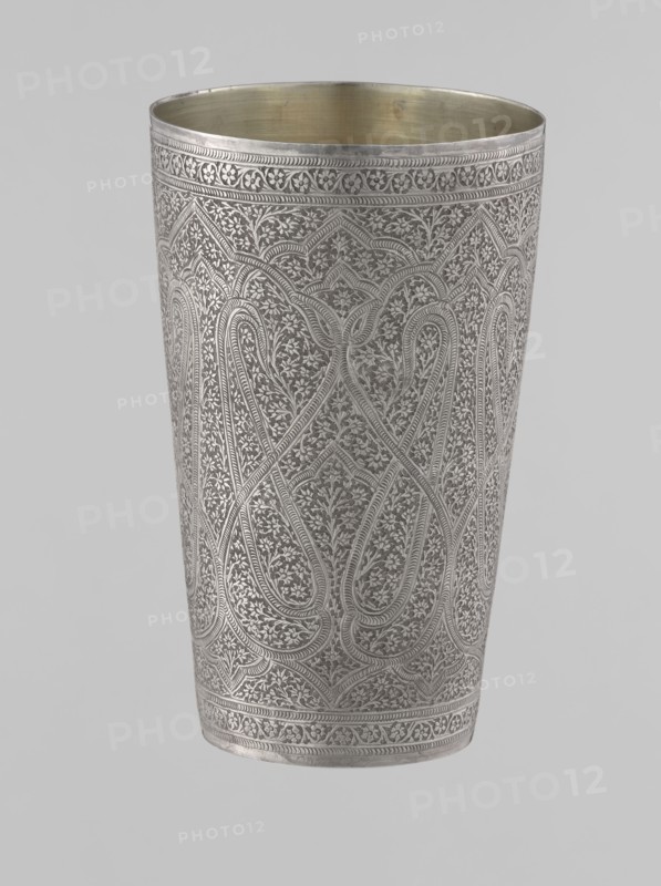 Mug, Islamic Mughal, Indian; 19th century, North India probable. Silver mug densely decorated with plant and floral motifs in a horror vacui style. Large symmetrical lines on the side wall form shapes reminiscent of flower buds; upper and lower rims decorated with horizontal ornamental lines. Interior gilded and polished. Vessel; weight 190 g; height 12.8 cm; diameter 7.8 cm., A tall metal cup with intricate fine engraving covering its exterior, showing repeating curving and floral motifs and bordered by decorative bands at the top and bottom; the exterior surface appears silver-gray while the interior shows a brass-toned golden color, and the background is a pale light gray.