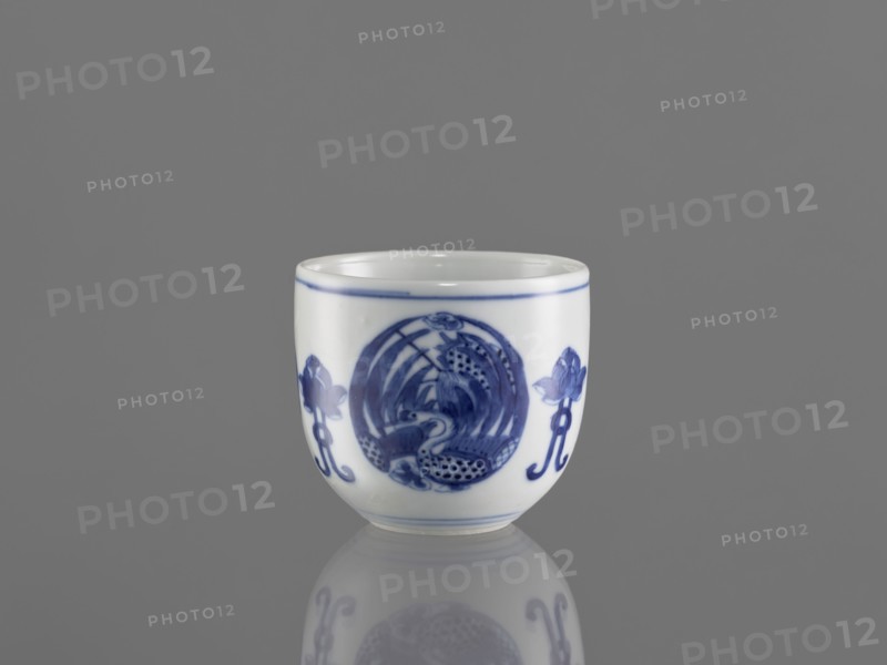 Bowl, ceramics, Chenghua 1465–1487, phoenixes and lotus iconography, blue-and-white porcelain in Qing style, floral ornaments, Bowl decorated with three phoenix images and stylized lotus flowers, early 20th century Qing dynasty, 1644–1912, China, vessel, height 7.5 cm, diameter 8 cm., A small white ceramic cup with blue painted decorative motifs sits centered on a reflective gray surface against a gray background; the cup shows a band of blue near the rim and a blue circular design on its front flanked by floral-like blue elements, and the surface reflection beneath the cup mirrors its shape and blue patterns.