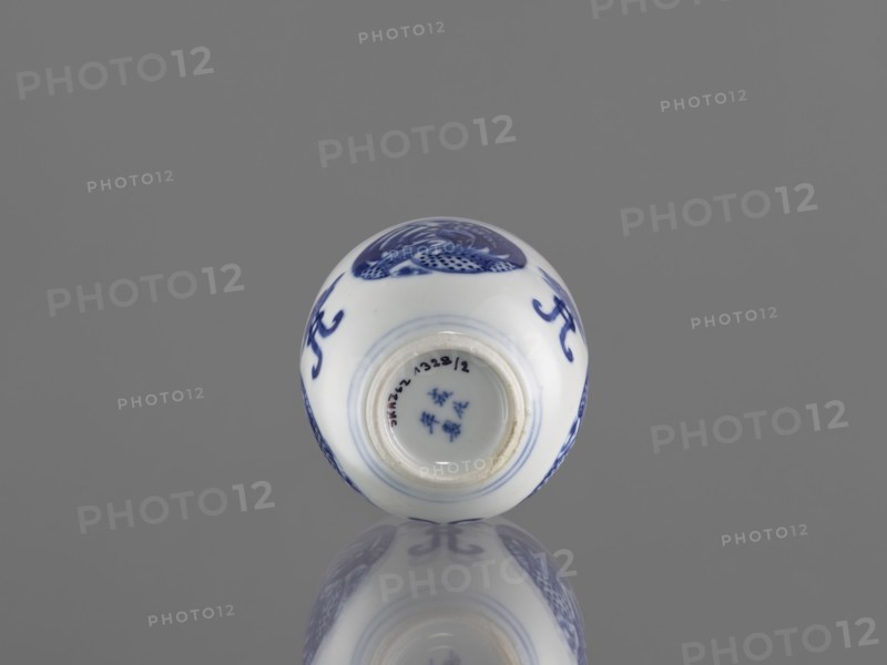 Bowl, ceramics, Chenghua 1465–1487, phoenixes, lotuses, blue-and-white porcelain, Qing style, Chinese art/culture, floral ornaments. Bowl decorated with three phoenix images and stylized lotus flowers. Early 20th century Qing dynasty, 1644–1912. China. Vessel; height 7.5 cm, diameter 8 cm., A small round ceramic object rests on a reflective surface against a solid gray background, showing white porcelain with blue painted decorative motifs and a glossy finish that creates a mirrored reflection below; the visible painted areas include curved brushstrokes and patterned designs in blue, and the rim of an opening at the top reveals the same white interior with blue detailing.