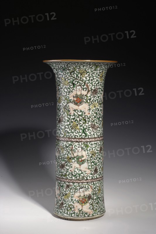 Cylindrical horn vase, Chinese, Qing dynasty, Kangxi period 1662–1722, colored porcelain in the famille verte palette. Ceramic vessel decorated with lotus tendril ornament and belt decoration. Height 37.5 cm; diameter 18.0 cm; weight 2.35 kg., A tall flared ceramic vase standing on a smooth surface, decorated all over with an intricate scrolling vine pattern in green, set against a white background with accents of brown, yellow, orange, and pale flesh tones; the surface shows a repeating motif of three seated figures positioned within the vinework, each figure wearing a green garment and holding a round orange object, and the vase is divided visually by a few narrow horizontal decorative bands in brown.