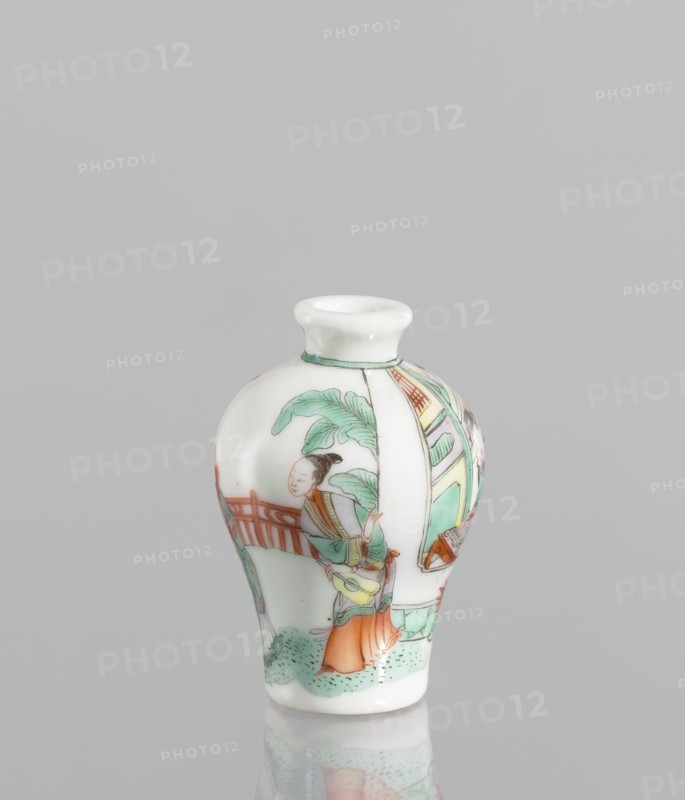 Flask - Snuffbox. A baluster-shaped flask, significantly widened at the shoulders, decorated with a fragment of landscape and architecture slightly highlighted in the decoration. Thick white porcelain body. 18th century, China. Colored porcelain, figurative scenes, images of women. Vessel snuffbox, height 7 cm, diameter 4.7 cm., A small porcelain vase with a rounded body and narrow neck sits against a plain light gray background; the vase is predominantly white with painted scenes in green, orange, red, light blue, brown, and black, showing a figure wearing orange and green garments standing near a railing and foliage rendered in green, with architectural elements in green and red on the curved surface, and the glaze gives a glossy finish with a faint reflection visible beneath the vase.