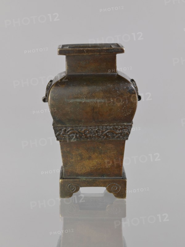 Fanggu vessel, Chinese, fanggu vessel, dragons iconography, Chinese art, ceremonial vessel, Qing 1644–1911, beasts fantasy, meander ornament, backdated signatures, 19th century, China, vessel, height 13.4 cm, width 9.0 cm, depth 7.9 cm, A small metal vessel with a square opening and a tapered body sits upright against a neutral gray background, showing an aged patina of brown, dark bronze, greenish and black tones; the upper section is squarish and the midsection bulges slightly with rounded shoulders and small ring-like handles on the sides, while a decorative band with raised carved motifs encircles the lower part of the bulge; the lower body narrows into a pedestal base that rests on four bracket feet with curled scrolled details, and the object casts a soft reflection on the glossy surface beneath it.