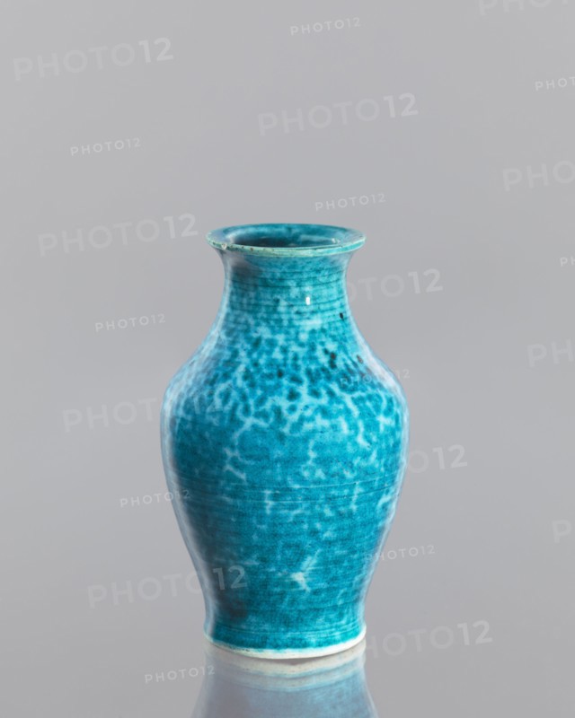 Vase, maker, Chinese, Qing dynasty 1644–1911, 2nd half of the 18th century, glazed crackleware ceramic vessel, height 12.5 cm, diameter 7.6 cm., A turquoise ceramic vase with a flared rim and a rounded body sits against a smooth light gray background, its surface showing mottled darker and lighter turquoise patterns and a glossy finish that produces subtle highlights and reflections, and a pale unglazed band is visible at the base where the vase meets a reflective surface that mirrors its shape.