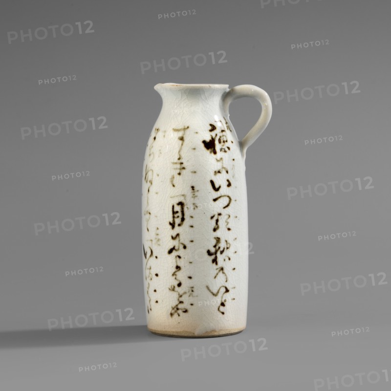 Cylinder-shaped jug with small handle and widened spout. White glaze with fine crackle; decoration painted underglaze with iron oxide black. Inscriptions; Japanese calligraphy. Ceramics, Japanese; 2nd half of the 19th century., Vessel, height 11.7 cm, width 6.3 cm, diameter 4.7 cm., A tall ceramic pitcher with a narrow, rounded body and a small curved handle sits against a plain gray background, the pitcher glazed in a pale off-white with a network of fine crackle lines visible on its surface; scattered across the body are several irregular dark brown brushlike marks and smudges that form vertical patterns, and the rim and base show subtle warm beige discoloration, while the handle matches the off-white glaze and the overall scene includes muted gray tones in the background and shadow.