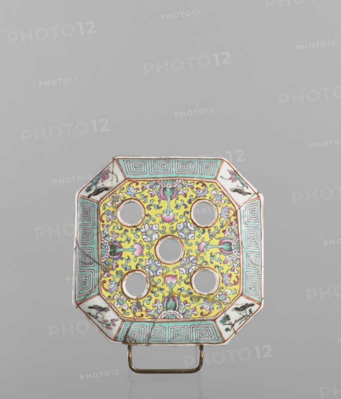 Vase with a lid, ceramics, colored porcelain, openwork perforated lid, decorated with flowers, birds, meander ornament, plant ornaments and lotus tendril motif. Flat, perforated lid. 19th century, China. Vessel. Height 27.0 cm; width 15.0 cm., An octagonal ceramic object with a central panel decorated in bright yellow and detailed with floral and scrolling motifs in pink, green, blue, and white, containing a grid of circular cutouts; the wide border features a repeating geometric pattern in turquoise and white with thin reddish outlines and small triangular corner panels painted with floral designs in pink, green, and black; the piece rests on a small metallic stand that is gold in color, and the background is a smooth neutral gray.