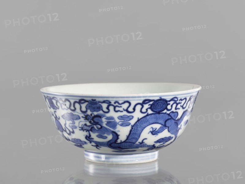 Bowl, ceramics with pearls, blue-and-white porcelain in Qianlong/Qing style, decorated with dragons and an ornamental band with eight Buddhist symbols at the top; Chinese, Qing dynasty Qianlong period, 1736–1795; vessel; height 5.5 cm, diameter 12.4 cm., A shallow ceramic bowl sits on a reflective surface against a plain gray background, showing a white interior and exterior decorated with a flowing blue painted design that includes a sinuous dragon motif, stylized cloud-like shapes, and ornamental borders; the rim and foot of the bowl have subtle blue accents, and the overall visible colors are white, various shades of blue, and gray.