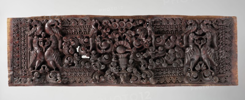 Decorative panel;, wooden panel likely originally a decorative element, part of home furniture or a door. The scene shows five pairs of birds—smaller parrots Skt. suka and larger geese Skt. hamsa—surrounded by lush plants, with a background of abundant floral and geometric ornament. Indian, South Asian; Indian art; birds; floral ornament; flowers; tree iconography. Sculpture, 18th century; height 24.0 cm, width 77.0 cm., A rectangular carved wooden panel with a dark brown to reddish-brown surface showing intricate raised relief work across its length; a pair of stylized bird-like creatures with curved necks and patterned bodies appear at each end, facing inward, and are flanked and connected by swirling vegetal scrolls and floral motifs; the central area features a clustered arrangement of carved rosettes and rounded blossom forms above a vase-like element, all surrounded by ornate lattice and repeated geometric border patterns; the wood shows lighter brown edges and small areas of wea...