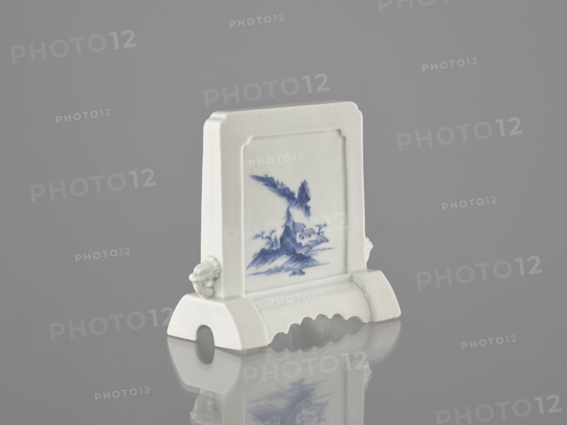 Decorative screen, ceramics; blue-and-white porcelain; Chinese, Qing dynasty 1644–1911; 19th century. Base cut with a wavy undercut; mascarons on the sides. Mountain landscapes depicted on both sides of the screen. Height 13.5 cm, width 13.7 cm., A small ceramic object stands on a reflective surface, predominantly white with blue painted decoration on its front panel; the piece has a rectangular upright section with a framed recessed panel showing a blue landscape motif, flanked by a pair of sculpted protrusions on the sides, and it rests on a curved base with an arched cutout and scalloped edge. The visible colors include white, blue, gray from the background, and subtle greenish tints on the glaze.