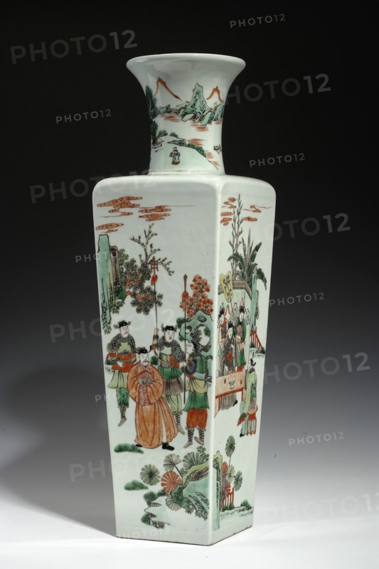Vase, Chinese; Qing dynasty 1644–1911, Kangxi period 1662–1722; porcelain, colored green family; cylindrical-neck vase; walls decorated with figural scenes depicting horsemen, courtiers, and women; symbolic ba bao emblems on the collar; mountain landscape with staffage on the neck; vessel; height 50.0 cm, width 14.7 cm, depth 14.7 cm., A tall, square-based porcelain vase with a flared neck stands against a dark background; the vase is predominantly white with painted scenes in green, orange, brown, black, and pale yellow. One side shows a group of figures in traditional clothing, with a central figure wearing an orange robe surrounded by attendants in green and brown garments, some holding long staffs; nearby are painted plants with green leaves and orange floral elements, stylized rocks, and wispy orange cloud motifs in the sky area. Another visible panel depicts additional figures seated and standing near a table and decorative screen, with green foliage and architectural elements in...