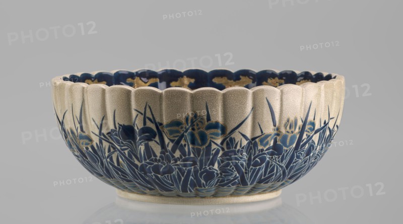 Bowl, Toyota Masanobu workshop, Satsuma ware, 2nd half of the 19th century Meiji period, Japan, Kagoshima. Deep bowl on a low ring-shaped base with a knobbed body and serrated rim, form reminiscent of a chrysanthemum. Outer wall decorated with clumps of blooming irises emerging from water; flowers and leaves painted in two-tone blue enamel, outlines and stylized waves/ripples in gold. Inner surface with a large gold chrysanthemum rosette imperial emblem in the mirror, Tokugawa family coat of arms inscribed in the center; side wall filled with dense, colorful ornament of various chrysanthemum flowers and butterflies in white, red, blue, gold, and green overglaze enamels and gold. Top edge finished with a uniform cobalt-blue valance. Signature on the base in gold reading: Satsuma yaki, Satsuma kuni, Ijun'in, Toyota Masanobu. Vessel height 12.0 cm, diameter 28.0 cm., A shallow scalloped ceramic bowl with a crackled off-white exterior rim and interior edge, decorated around its lower exter...
