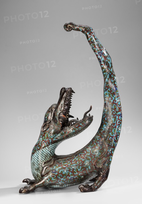 Dragon, gift, art of Southeast Asia, Southeast Asia culture, images of animals, fantastic creatures, dragons, 19th century, Southeast Asia, sculpture, height 51.0 cm, width 35.0 cm, A sculptural creature with an elongated curved body and upturned tail, shown in a three-quarter view, rendered in metal with intricate surface patterns in turquoise, blue, green, brown, and reddish tones; it has an open mouth with visible teeth and a forked tongue, a textured throat and ribbed underside, small rounded ears, clawed feet, and scrolling decorative motifs covering its surface against a plain light gray background.