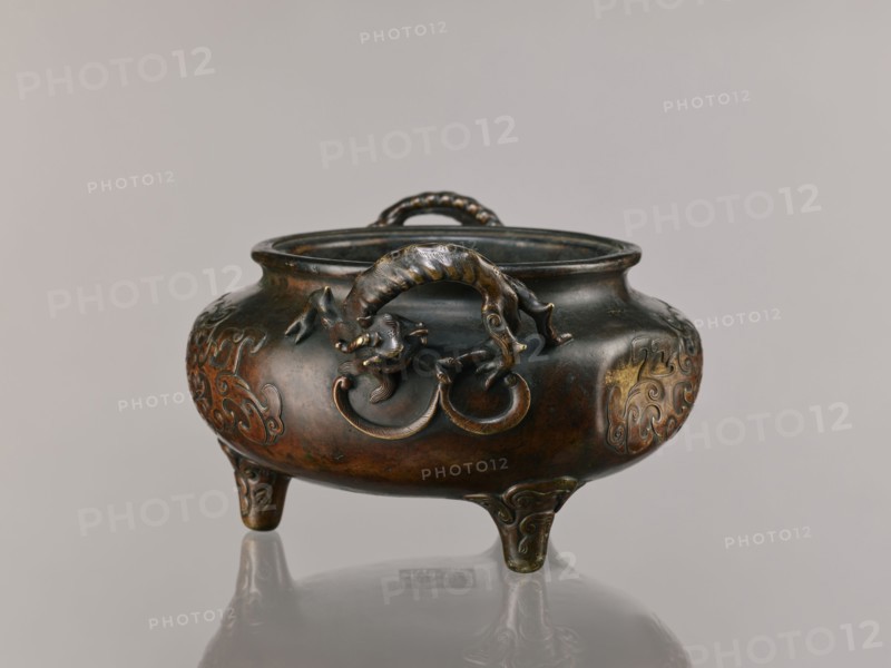 Ding incense burner, Chinese, Qing 1644–1911, backdated signatures, dragon-shaped handles. Based on ceramics from the Xuande era 1426–1435 of the Ming dynasty; 19th century, China. Ceremonial/ritual vessel with lizards, dragons, and taotie iconography. Height 13.1 cm; diameter 21.4 cm., A low, rounded tripod censer with a wide mouth and a curled handle on top, cast in a dark metallic material with polished highlights in brown, bronze, gold, and greenish patina; the body rests on three short curved legs and bears raised decorative motifs, while a sculpted mythical creature with a sinuous body and curled tail clings to the front edge holding a ring in its mouth, and the object sits on a reflective pale surface against a neutral light gray background.