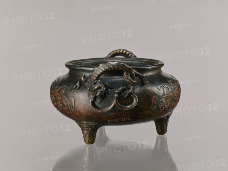 Ding incense burner, Chinese; Qing 1644–1911, 19th century, based on ceramics from the Xuande era 1426–1435 during the Ming dynasty. Bronze vessel, ceremonial/ritual incense burner with dragon-shaped handles; motifs include lizards, dragons, taotie; backdated signatures. Height 13.1 cm, diameter 21.4 cm., A rounded metal vessel with a dark brown and greenish patina rests on a reflective surface, supported by three short ornate legs; the body shows raised swirling decorative motifs and sculpted handles shaped like a curled animal with a textured body gripping circular rings, and a braided loop handle spans the rim; visible colors include dark brown, greenish bronze, and hints of golden wear.