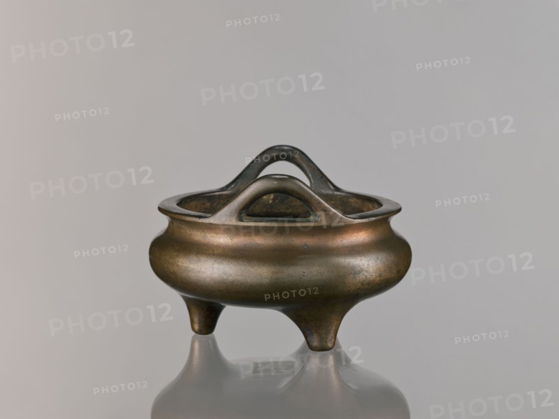 Li-type incense burner, Chinese, Qing dynasty 1644–1911, 18th century. Round, flattened Li-type ritual/ceremonial incense burner on three low legs, with backdated signatures. Height 10.0 cm, diameter 15.7 cm., A small rounded metal vessel with a smooth, slightly bulbous body and three short tapering legs, shown against a plain light gray background; the vessel has an open top with an integrated looped handle structure that rises above the rim in a double-arched shape, and the metal surface shows a patina with shades of bronze, brown, muted gold, and hints of darker gray, with subtle areas of wear and slight greenish spots; the vessel casts a soft reflection on the glossy surface beneath it.