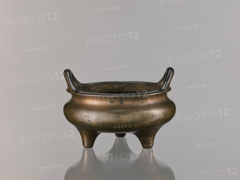Li-type incense burner, Qing dynasty 1644–1911, 18th century, Chinese, round flattened form on three low legs, Li-type/ritual/ceremonial vessel, incense burner, with backdated signatures, height 10.0 cm, diameter 15.7 cm, A small metal vessel with a rounded body and a wide rim sits on a reflective surface, supported by a group of three short, tapered legs; the rim rises into a pair of upward-curving handles on opposite sides. The vessel shows a patina with shades of brown, bronze, dark greenish spots, and darker brown to almost black areas inside the rim and on the handles. The background is a smooth, even light gray, and the reflection beneath the vessel reproduces its colors and shape faintly.