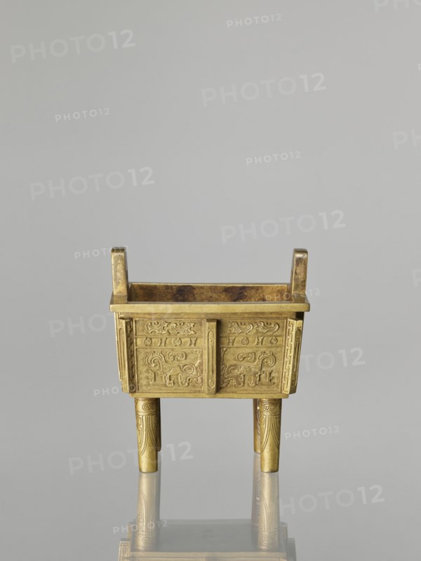 Fangding incense burner with cover, Chinese, Qing dynasty 1644–1911, 19th century. Bronze vessel decorated with archaic ornament in flat relief, featuring taotie masks and fantastic creatures. Height 15.2 cm, width 14.0 cm, depth 10.9 cm., A small rectangular metal vessel with a gold color sits on four cylindrical legs and has two short vertical handles at the top corners; the body of the vessel is decorated with intricate raised patterns and swirling motifs on all visible sides, and the legs show linear carved designs; the interior of the vessel has a darker, slightly mottled gold-brown tone, and the whole object casts a soft reflection on the pale gray surface beneath it; visible colors include gold, darker gold-brown, pale gray, and subtle shadowed grays.