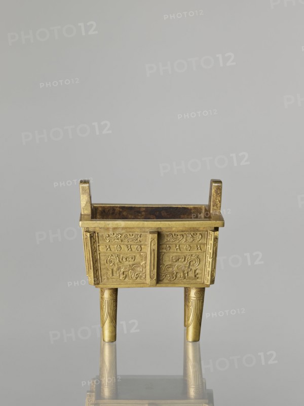 Fangding incense burner with cover, Chinese, Qing dynasty 1644–1911, 19th century. Bronze vessel decorated on the walls with archaic ornament in flat relief, taotie and fantastic creature motifs; ceremonial vessel. Height 15.2 cm, width 14.0 cm, depth 10.9 cm., A small rectangular metal vessel with a golden color sits on three tapered legs and has two short upright handles at opposite ends; the vessel's sides feature raised decorative patterns with curving motifs and panels, and the legs show incised linear designs; the interior appears darker with a rich brown tone, and the object casts a subtle reflection on a smooth gray surface beneath it against a pale gray background.