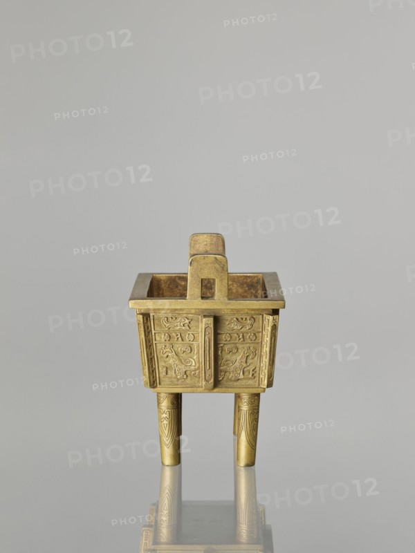 Fangding incense burner with cover; Chinese; Qing dynasty 1644–1911, 19th century; decorated on the walls with archaic ornament in flat relief and taotie/ fantastic creatures; ceremonial vessel; materials/technique: Chinese decorations; height 15.2 cm, width 14.0 cm, depth 10.9 cm., A small metallic vessel sits centered against a smooth light gray background, its surface showing a warm golden color with subtle darker patina in places. The vessel has a square top opening with a flat rim and a rectangular handle rising from the center, both matching the golden tone. The body tapers slightly toward the base and displays raised decorative patterns on its sides in the same golden color with darker shading accentuating the designs. The vessel stands on three cylindrical legs, also golden, each engraved with simple vertical motifs, and a faint reflection of the vessel appears on the glossy surface beneath it.
