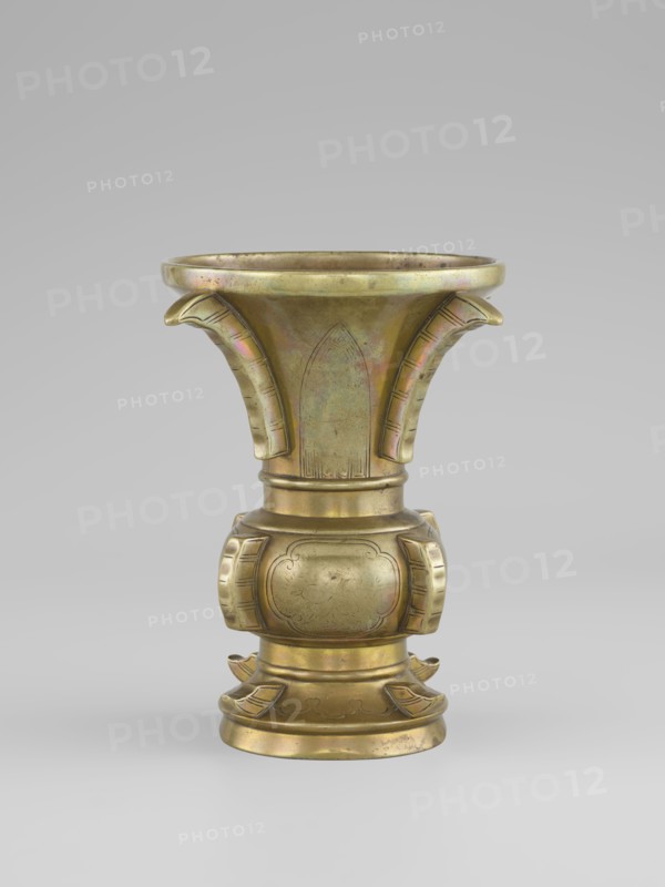 Gu-type vase, Chinese, Qing dynasty 1644–1911, 19th century. Ritual/ceremonial vessel inspired by the cult gu shape with four convex, interrupted strips. Decoration: on the belly, oval reserves with stylized peonies on a grained background. Porcelain/ceramic vessel, height 16 cm, diameter 12.2 cm., A single metallic vessel stands centered against a neutral gray background, displaying a warm golden-bronze color with subtle iridescent patches of green and pink on its surface. The vessel has a flared, trumpet-like upper section with a rounded rim and two curved, stylized protrusions on opposite sides, each showing engraved linear details. The midsection tapers into a rounded, bulbous body decorated with two symmetrical framed panels and faint engraved motifs. The lower portion rests on a stepped, circular base accented by outward-curving, petal-like projections and engraved decorative lines, all sharing the same golden-bronze tone with spots of darker patina.