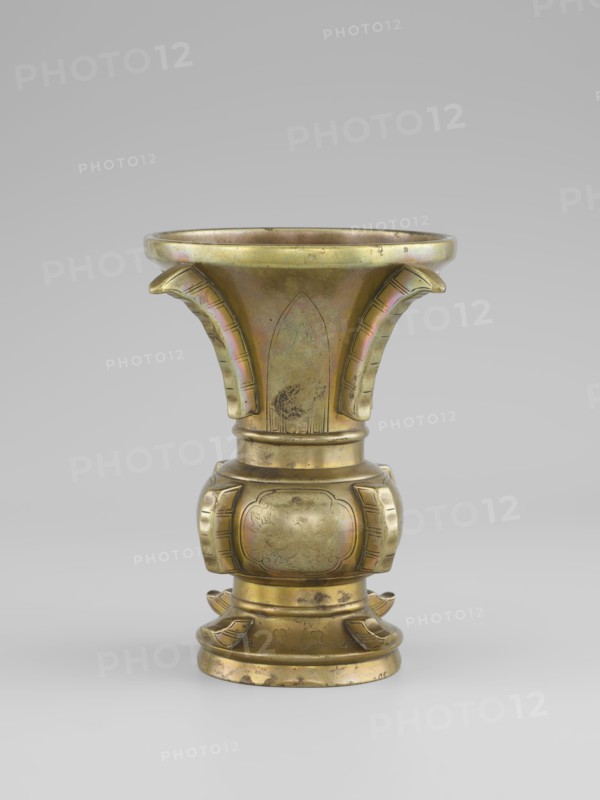 Gu-type vase, Vase with a shape inspired by the cult vessel gu, with four convex, interrupted strips. Decoration: on the belly in oval reserves stylized peonies on a grained background. Ceremonial/ritual vessel, Qing dynasty 1644–1911, 19th century China. Porcelain/ceramic vessel, height 16 cm, diameter 12.2 cm., A metallic vessel with a flared rim and a rounded base, displaying a warm golden color with areas of greenish and purplish iridescence and darker tarnished spots; the upper section flares outward with three curved, ribbed projections extending downward, each showing subtle linear incisions; the midsection narrows into a cylindrical neck that leads to a bulbous, paneled body decorated with shallow carved motifs and recessed rectangular panels on its sides, all in the same golden tone with slightly darker engraved lines; the lower section steps down into a circular foot with layered rings and small curved adornments around its perimeter, showing patches of wear and darker discol...