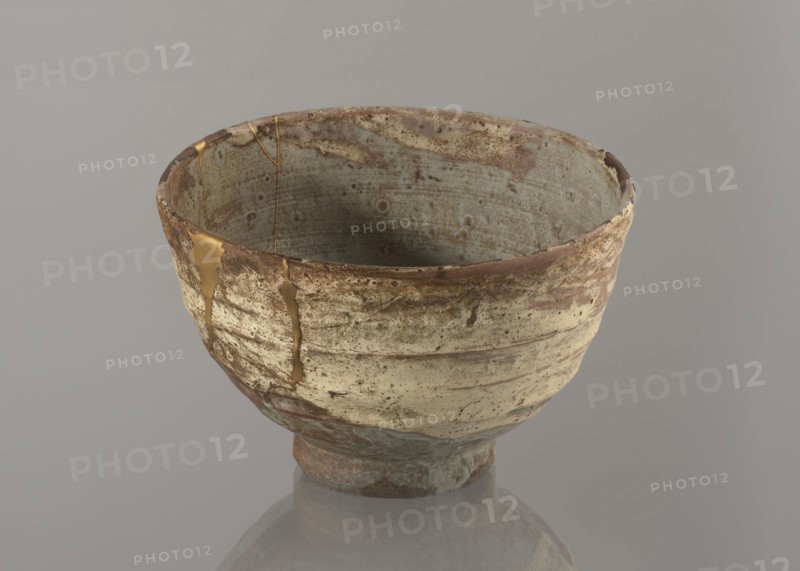 Wan-nari type tea bowl wan-nari chawan, Japanese, Edo 1603–1868, 18th–19th century. Stoneware vessel on a low, round foot; rough surface; partly glazed with white milky and grey-bluish glaze with numerous spots and drips, gold lacquer veins. Fillings and gluing made with gold lacquer. Height 8.4 cm; diameter 13.5 cm., A ceramic bowl sits on a smooth surface against a neutral backdrop, showing an irregular, rounded shape with a slightly flared rim and a narrow foot; the bowl’s exterior and interior surfaces display a weathered texture with mottled patches and speckled markings. The visible colors include muted beige and cream on the exterior, pale blue-gray inside, brown and rusty tones around the rim and base, and a metallic gold filling along repaired cracks. The bowl’s surface shows horizontal striations and areas where glaze appears thin or worn, and the gold repairs create shiny, smooth lines contrasting with the rougher, matte clay. The neutral background is a soft gray, and a fai...