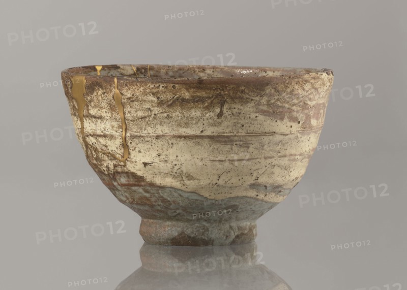 Wan-nari type tea bowl wan-nari chawan, noted. Japanese, Edo period 18th–19th century. Stoneware bowl on a low, round foot; rough surface; partly glazed with white milky and grey-bluish glaze with numerous spots and drips and gold lacquer veins. Fillings and gluing made with gold lacquer. Function: tea vessel chawan. Height 8.4 cm; diameter 13.5 cm., A ceramic bowl with a worn, textured surface sits against a plain background, showing a palette of beige, brown, tan, gray, blue-gray, and gold. The bowl has an uneven rim and mottled areas where darker brown and rust tones blend into lighter beige and tan, with speckled dark spots and subtle horizontal striations. A few gold-colored repairs trace irregular lines along the side and rim. The lower portion shifts to cooler gray and blue-gray hues with rougher texture, and the bowl rests on a short, slightly irregular foot that mirrors the same earthy tones. The background is a smooth neutral gray.