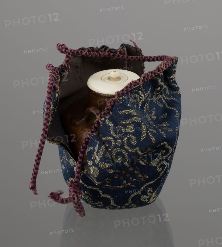 Tea canister chaire of the mimitsuki type. Brown container with two handles and a lid in a navy blue brocade bag silk. Dark brown, porous clay, wheel-thrown; light brown glaze with rough air bubble cracks; applied plastic handles. 19th century late Edo–Meiji period, Owari, Japan. Height 6.3 cm; diameter 5 cm. Function: tea container chaire., A fabric pouch with a drawstring closure sits open to reveal a rounded container inside; the pouch fabric is predominantly deep blue with a metallic gold floral and scroll pattern and has a dark interior lining, while the drawstring is a braided purple cord with frayed ends; the container inside appears brown with a glossy surface and a creamy off-white lid with a small raised knob; the background is a smooth neutral gray and the objects cast a subtle reflection on the surface below.