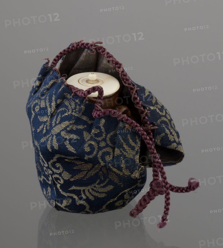 Tea canister chaire of the mimitsuki type, Seto; Japanese, Edo 1603–1868 / Meiji 1868–1912, 19th century; brown container with two handles and a lid in a navy blue silk brocade bag; dark brown porous wheel-thrown clay with light brown glaze showing rough air-bubble cracks; plastic handles; height 6.3 cm, diameter 5 cm; Owari, Japan., A fabric pouch with a blue background patterned in gold sits open, revealing a light-colored circular lid and a glimpse of a brown rounded object inside; the pouch has braided drawstring cords in a purple tone that form a knot and hang down, and the pouch interior appears darker, while the overall scene rests against a neutral gray surface and background.