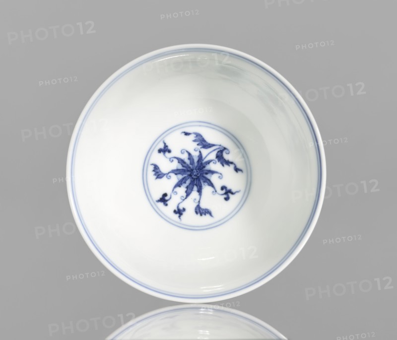 Bowl, blue and white porcelain, Chinese, Qianlong style, Qing dynasty 1644–1911, 1736–1795. Porcelain vessel with lotus and lotus tendril ornament; slightly flared bowl. Height 4.3 cm; diameter 9.8 cm., A white ceramic bowl with a smooth glossy surface sits against a gray background, showing a delicate blue floral-like motif painted at the center inside a thin blue circular border, and thin blue lines rim the inner edge; the decoration includes flowing petal and vine shapes in blue, and the bowl's interior and exterior appear predominantly white with highlights and reflections of light, while the background is gray.