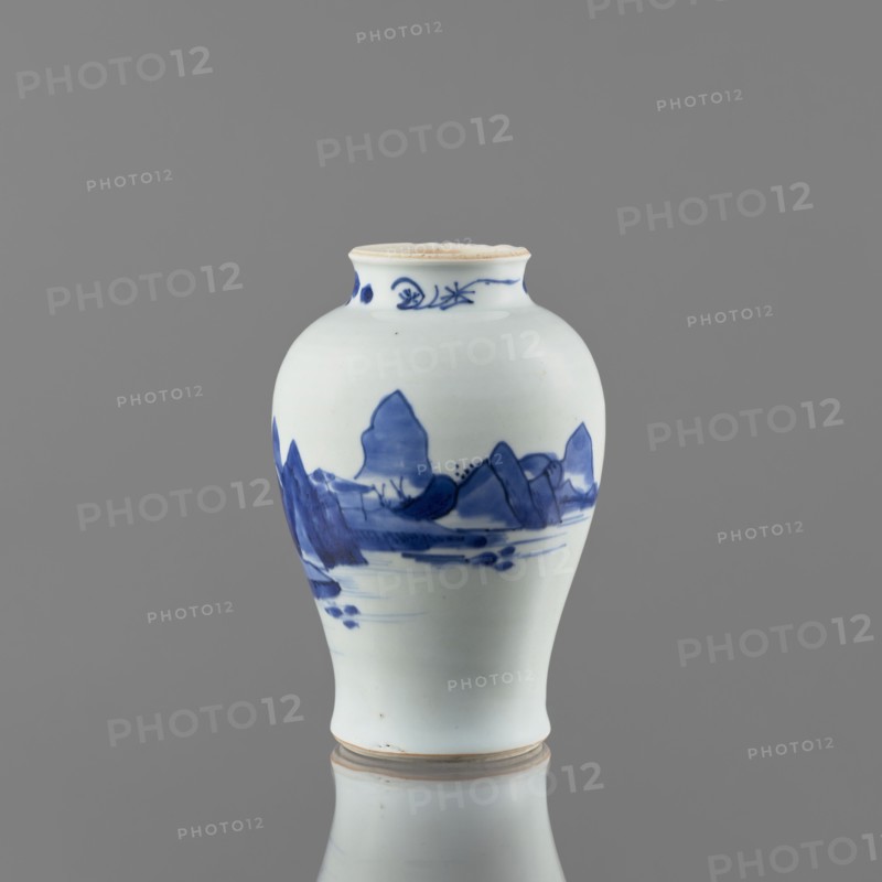 Meiping-type vase, ceramics, Meiping vessel type, blue and white porcelain, Qing style, 1662–1722, Chinese, decorated with images of rocks, trees, and a pagoda; neck decorated with small plant motifs; short neck, height 17.7 cm, diameter 11.8 cm, made in China., A small rounded ceramic vase with a narrow neck and slightly flared rim sits against a plain gray background, predominantly white in color with blue painted landscape motifs wrapping around its midsection; the blue decoration shows mountain-like shapes, horizontal brushstrokes suggesting water, and a few darker spots, while the rim and base have a light beige edge and the vase is reflected faintly below, showing the same white, blue, and beige tones.