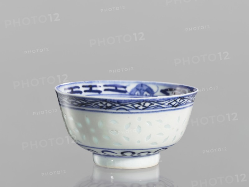 Bowl, Chinese, Qing 1644–1911, ceramics; blue-and-white porcelain with rice-pattern and geometric and plant/ floral lotus decorations. Bowl on a low ring foot, decorated on the body with a rice grain ornament and geometric-plant borders; in the mirror a lotus flower in a double circle. 18th–19th century, China; vessel; height 5.5 cm, diameter 10.3 cm., A ceramic bowl with a glossy white surface and blue decorative patterns around the rim and base sits on a reflective surface; the rim features a darker blue band with geometric motifs and a thinner blue line below it, the interior rim also shows blue designs, and the bowl’s body displays a subtle pattern of small, pale leaf-like shapes against the white glaze, with the visible colors being white, several shades of blue, and a hint of pale green from reflections, all set against a plain light gray background.
