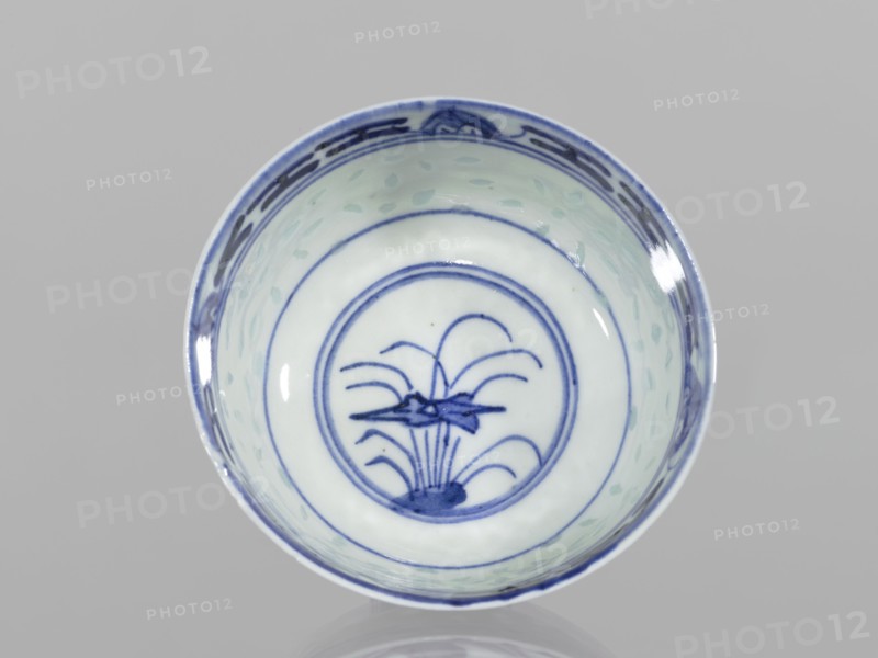 Bowl, Chinese, Qing dynasty 1644–1911, 18th–19th century. Blue-and-white porcelain, rice pattern with geometric and plant ornaments and floral decorations including lotuses. Bowl on a low ring foot, decorated on the body with a rice grain ornament and geometric-plant borders; in the mirror a lotus flower in a double circle. Vessel; height 5.5 cm, diameter 10.3 cm., A round ceramic bowl viewed from above with a pale white interior and blue painted decoration, showing a central circular motif with stylized plant-like lines and curved shapes in blue, concentric blue rings around the center, a patterned blue band near the rim, and a few light green brushstroke spots on the inner walls; the rim and painted details appear in shades of blue against the white background, and the overall scene is set on a neutral gray surface.