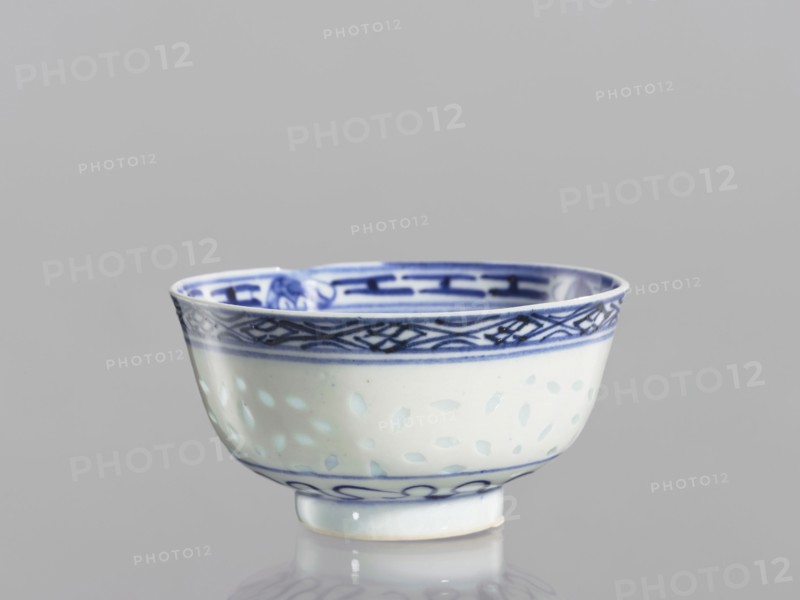 Bowl, Chinese, Qing dynasty 1644–1911, 18th–19th century. Blue-and-white porcelain bowl on a low ring foot, height 5.3 cm, diameter 10.3 cm. Decorated on the body with a rice grain rice pattern ornament, geometric and plant borders, floral decorations including lotuses; in the mirror a lotus flower in a double circle., A small porcelain bowl with a rounded shape and a short foot, predominantly white with blue decorative patterns; the rim and inner band feature darker blue geometric and interlaced designs, while the exterior body shows a softer blue dotted or leaf-like motif and a thin blue line near the base; the bowl sits on a reflective surface and the background is a smooth light gray.