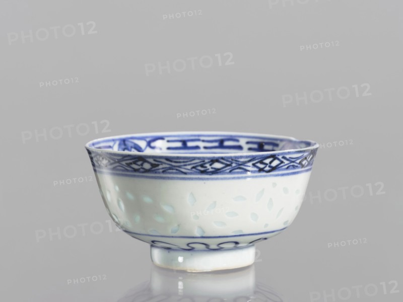 Bowl, unknown provenance; Chinese; Qing dynasty 1644–1911; blue-and-white porcelain; 18th–19th century; vessel on a low ring foot, height 5.3 cm, diameter 10.3 cm; decorated on the body with a rice grain pattern, geometric and plant borders; mirror with a lotus flower in a double circle; motifs include rice pattern, geometric ornaments, plant ornaments, floral decorations, lotuses., A small ceramic bowl sits on a reflective surface against a plain gray background, showing white and blue as the dominant colors; the bowl has a white body with blue decorative bands near the rim and base, a patterned blue border around the upper interior and exterior, and a subtle light blue leaf-like motif scattered on the midsection; the rim is slightly undulating and the bowl rests on a short circular foot with a blue line near its top, and soft reflections and highlights are visible on the glazed surface.