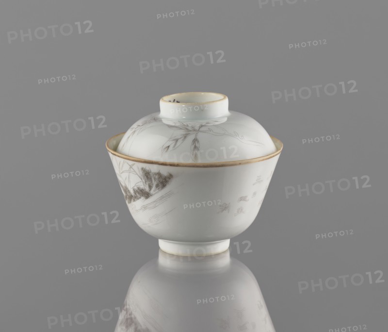 Bowl with a lid, decorated with ducks and peaches, colored porcelain. Chinese, Qing dynasty, Tongzhi period 1862–1874. Porcelain bowl on a ring foot with a lid-stand; exterior painted with a duck among reeds in delicate black line and an inscription. Lid decorated with a branch bearing peach fruit. Vessel, height 9 cm, diameter 10.3 cm., A white porcelain lidded cup with a shallow domed lid sits centered on a reflective surface against a gray background; the cup and lid have thin gold rims and delicate grayish-brown painted botanical and landscape motifs including slender leaves and a textured cluster resembling foliage, and the cup shows faint decorative marks in the same grayish-brown tone, with the reflection of the cup visible below.