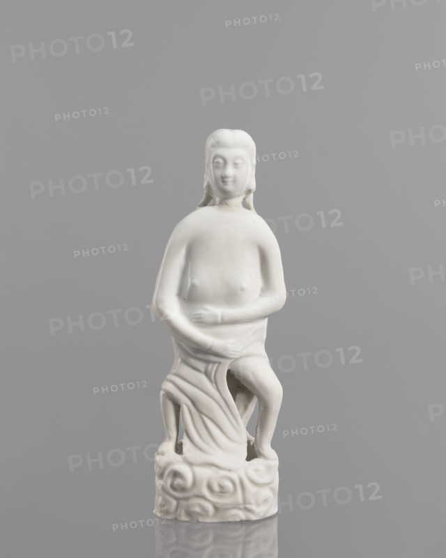 Adam figurine, Chinese, 17th/18th century. Sculpture of a half-naked Adam Biblical, male figurine; height 20.3 cm, width 7.0 cm., A smooth white ceramic figurine of a seated person with long hair, a calm facial expression, and bare upper torso, shown against a uniform grey background; the figure sits on a decorative base with rounded, swirling shapes and draped cloth over the lower body, with one leg bent and visible, and the surface reflecting subtle highlights.