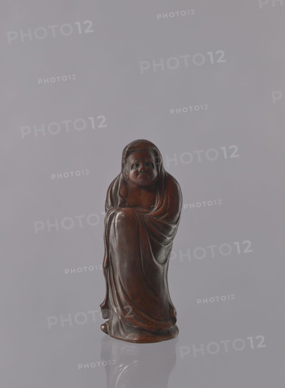 Daruma, unknown sculptor, Japanese, Meiji period 1868–1912, ceramics figurine of a male Buddhist monk Daruma wearing headgear and a long, flowing robe covering the head, late 19th century, Seto, Japan; height 21.2 cm, A small carved figure made of dark brown material stands centered against a smooth light gray background, showing a robed person with a rounded head and a solemn facial expression, draped garments with flowing folds that wrap around the body, subtle highlights and deeper brown shadows on the surface, and a glossy finish that creates a faint reflection beneath the base.