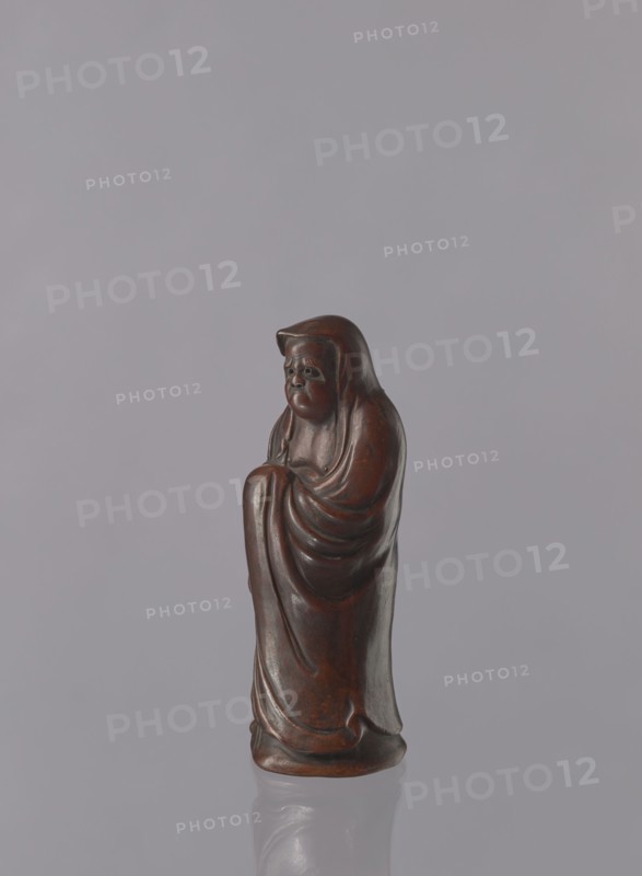 Daruma, Japanese, Meiji period 1868–1912. Ceramic figurine of Daruma Buddhist monk, male figure in a long flowing robe covering the head and headgear. Late 19th century, Seto, Japan. Height: 21.2 cm., A small carved figurine with a rounded head and draped cloak, standing upright on a smooth base, showing curved folds and rounded contours; the surface is polished and appears dark brown with lighter brown highlights, set against a pale gray background with a subtle reflection of the base on a glossy surface.